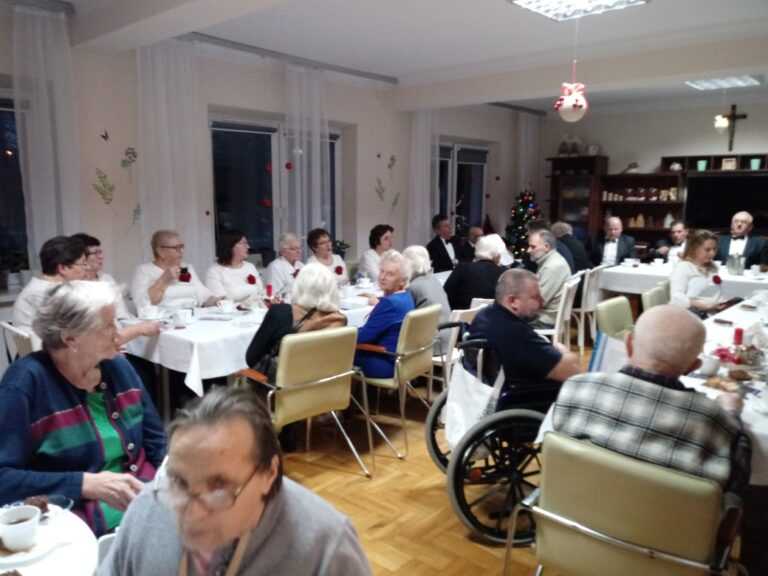 mieszkańcy DPS i chórzyści siedzą przy stołach, częstują się przygotowanym poczęstunkiem i rozmawiają