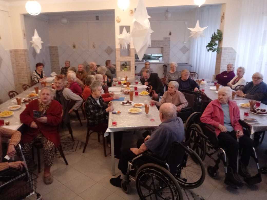 mieszkańcy DPS siedzą przy stołach, częstują się przygotowanym poczęstunkiem i śpiewają kolędy, wyświetlane przez rzutnik multimedialny