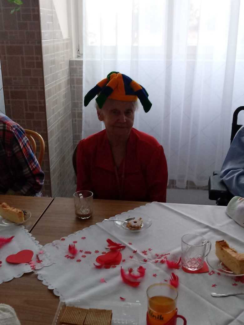 mieszkanka DPS podczas zabawy walentynkowej siedzi za stołem w śmiesznym nakryciu głowy i uśmiecha się