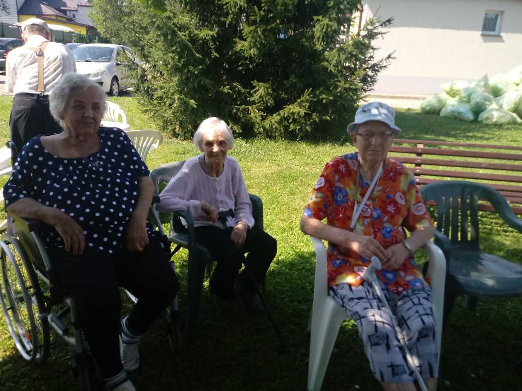 mieszkańcy biorący udział w grillu w ogrodzie DPS siedzą na krzesłach w cieniu drzew