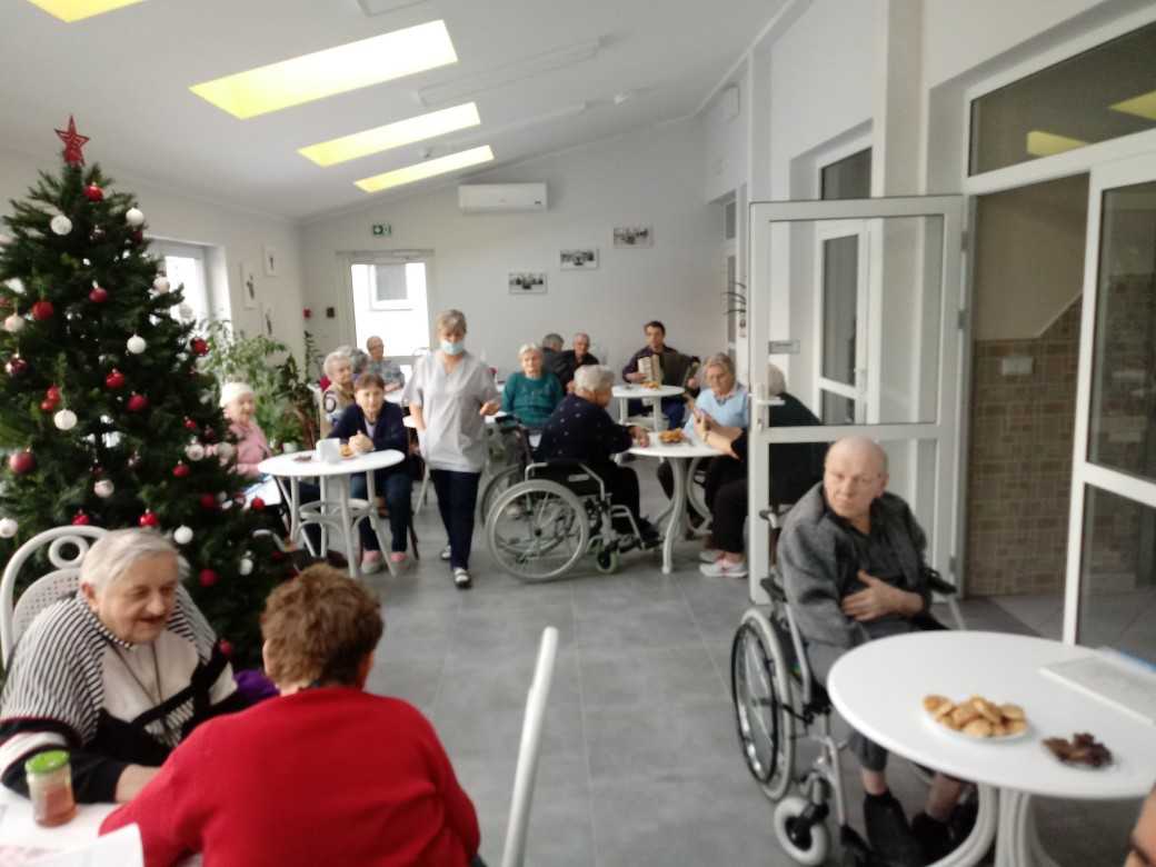 mieszkańcy znajdują się w ogrodzie zimowym, siedzą przy stolikach, rozmawiają, częstują się przekąskami, śpiewają kolędy