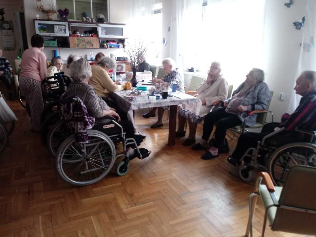 mieszkańcy DPS siedzą wokół stołu na świetlicy, rozmawiają i wykonują prace manualne w ramach terapii zajęciowej