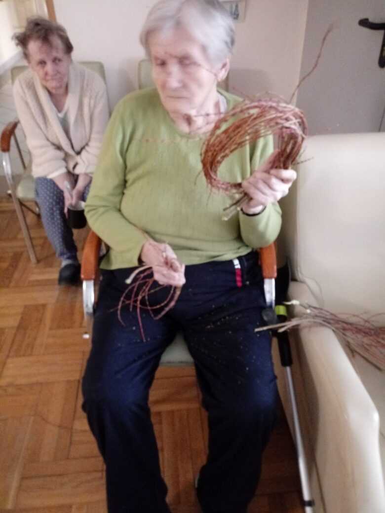 mieszkanka DPS siedzi na fotelu i plecie wianek z wikliny
