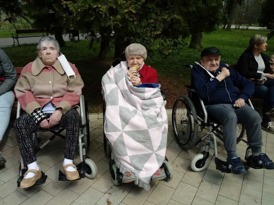 mieszkańcy DPS siedzą na wózkach inwalidzkich w parku i jedzą lody