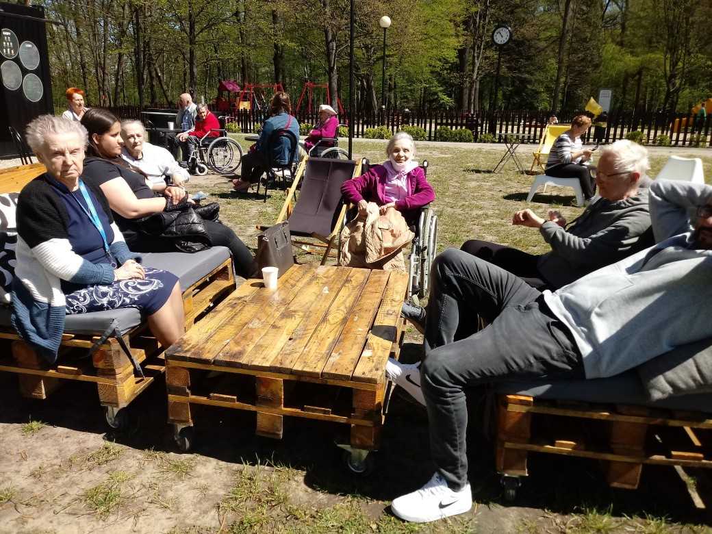 mieszkańcy i pracownicy DPS spędzają czas w parkowej kawiarence na świeżym powietrzu, siedzą przy stoliku, rozmawiają, wygrzewają się w promieniach słońca