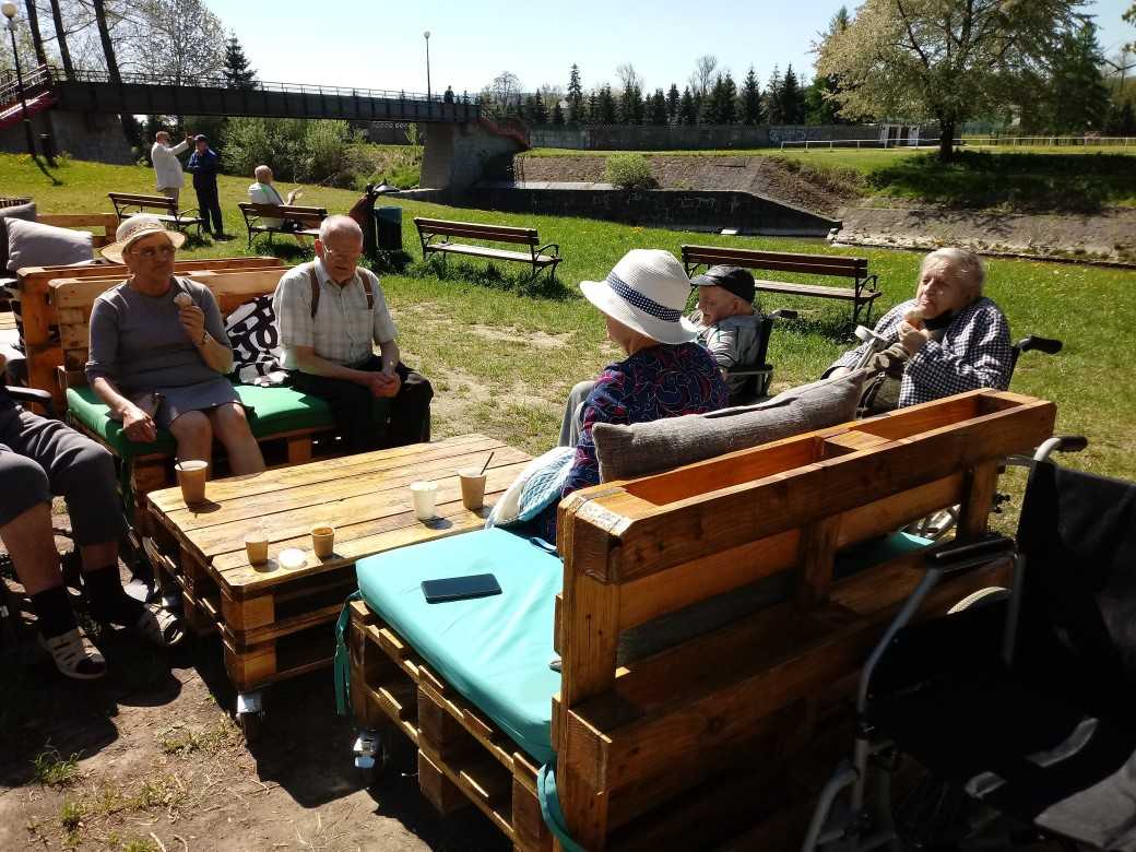 mieszkańcy DPS siedzą przy stoliku w parku nad rzeką, jedzą lody, rozmawiają