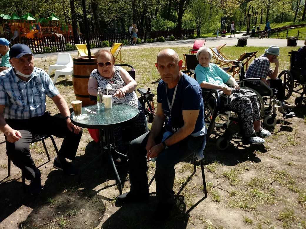 mieszkańcy DPS siedzą przy stoliku w parkowej kawiarence na świeżym powietrzu, piją kawę i wodę