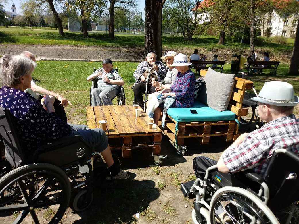 mieszkańcy DPS spędzają czas w parku nad rzeką, siedzą przy stoliku, jedzą lody, piją kawę, rozmawiają, ogrzewają się w promieniach słońca