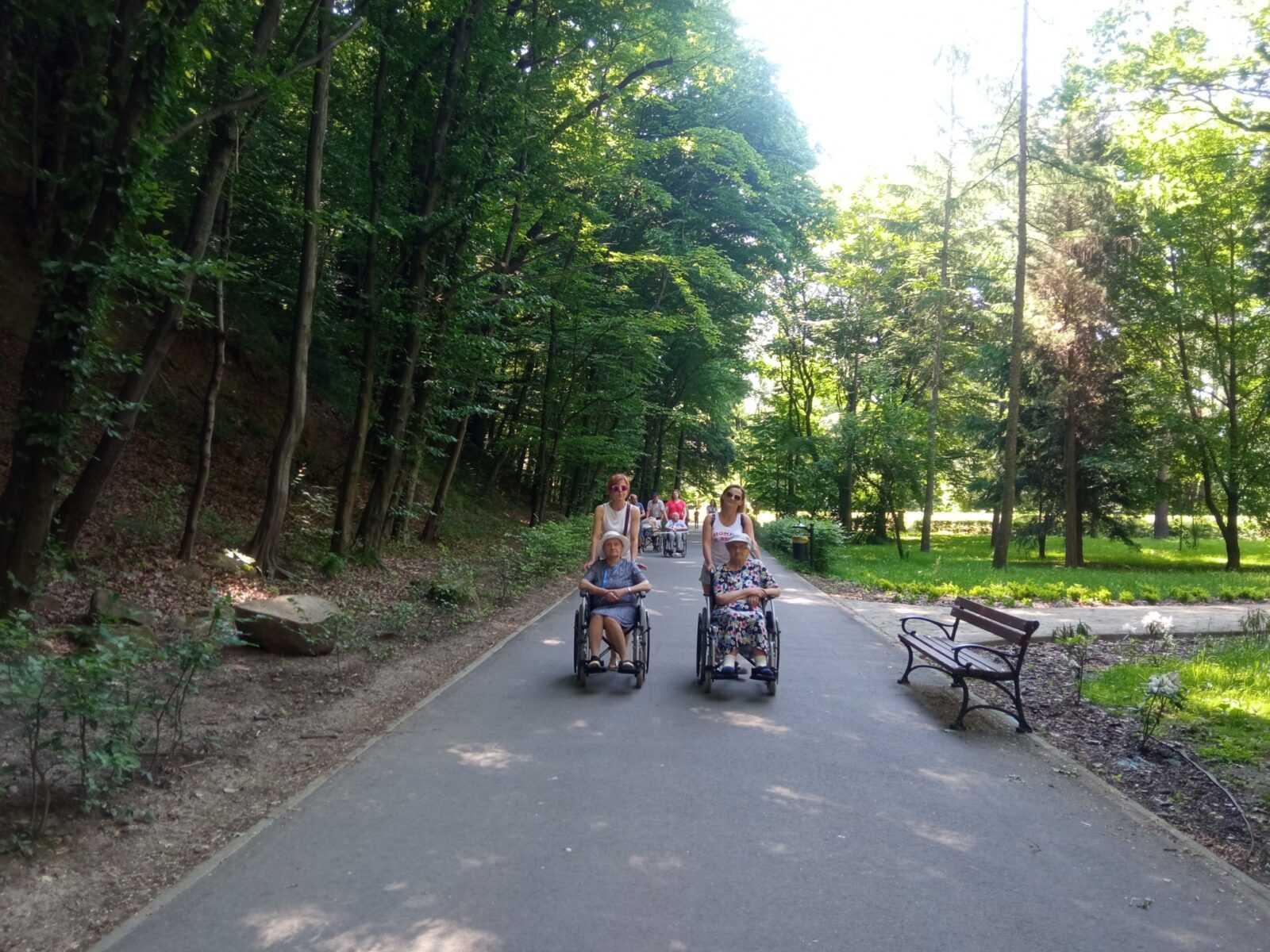 podczas spaceru po parku pracownice DPS pchają alejką wózki inwalidzkie, na których siedzą mieszkańcy DPS
