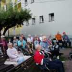 grupa starszych ludzi siedzi na włkach i wózkach inwalidzkich w ogrodzie i ogląda film