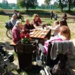 starsi ludzie siedzą przy stole w parku, rozmawiają, jedzą ciastka i piją kawę