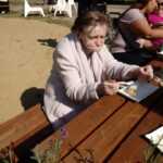 starsza pani siedzi na ławce w parku przy stoliku i je kremówkę