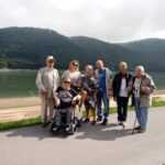 grupa osób starszych i niepełnosprawnych pozuje do zdjęcia na tle jeziora i gór